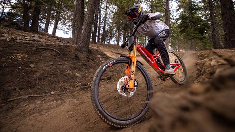 A mountain biker on a red Santa Cruz bike in black pants, a white jersey, and black full face helmet on a Summit Bike Park with trees lining the dirt trail.