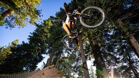 A mountain biker in khaki pants and a white tee shirt getting air on their bike off a paved dirt berm at Summit Bike Park with the sun setting through the trees in the background.