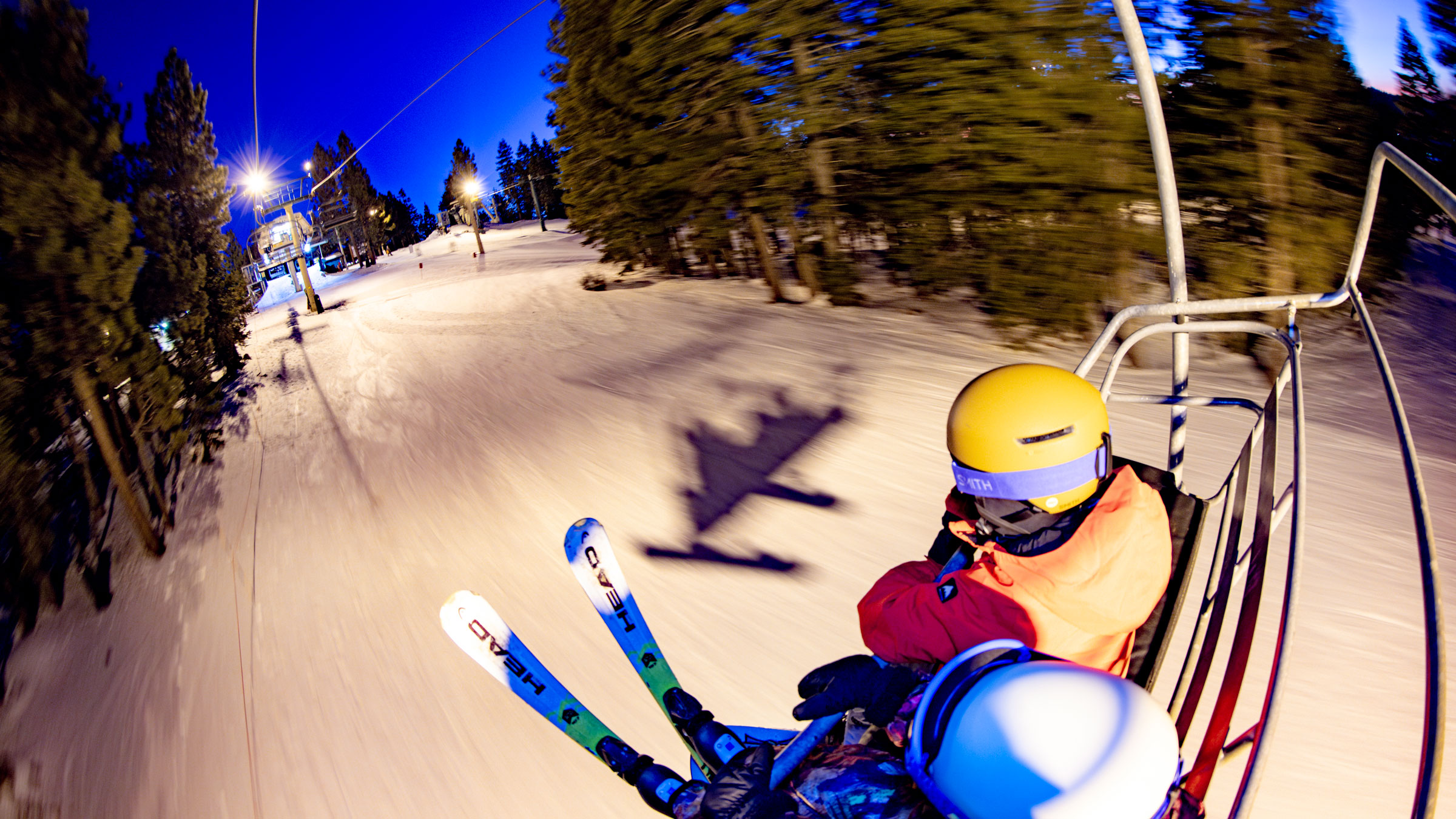 Two kiddos on a chairlift, one in a yellow helmet and the other in white helmet with white Head branded skis, during a Night Session at Snow Summit during winter.