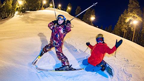 Two kiddos on a trail at Snow Summit during a winter evening Night Session, one in a colorful ski outfit on skis holding their ski poles up and the other in a red jacket on a snowboard.