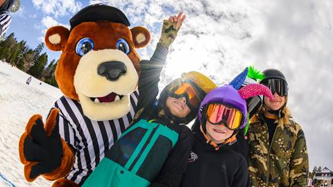 Big Bear Mountain Resort's mascot, Biggie the Bear, posing with three kiddos on the slopes of Bear Mountain during a sunny winter day.