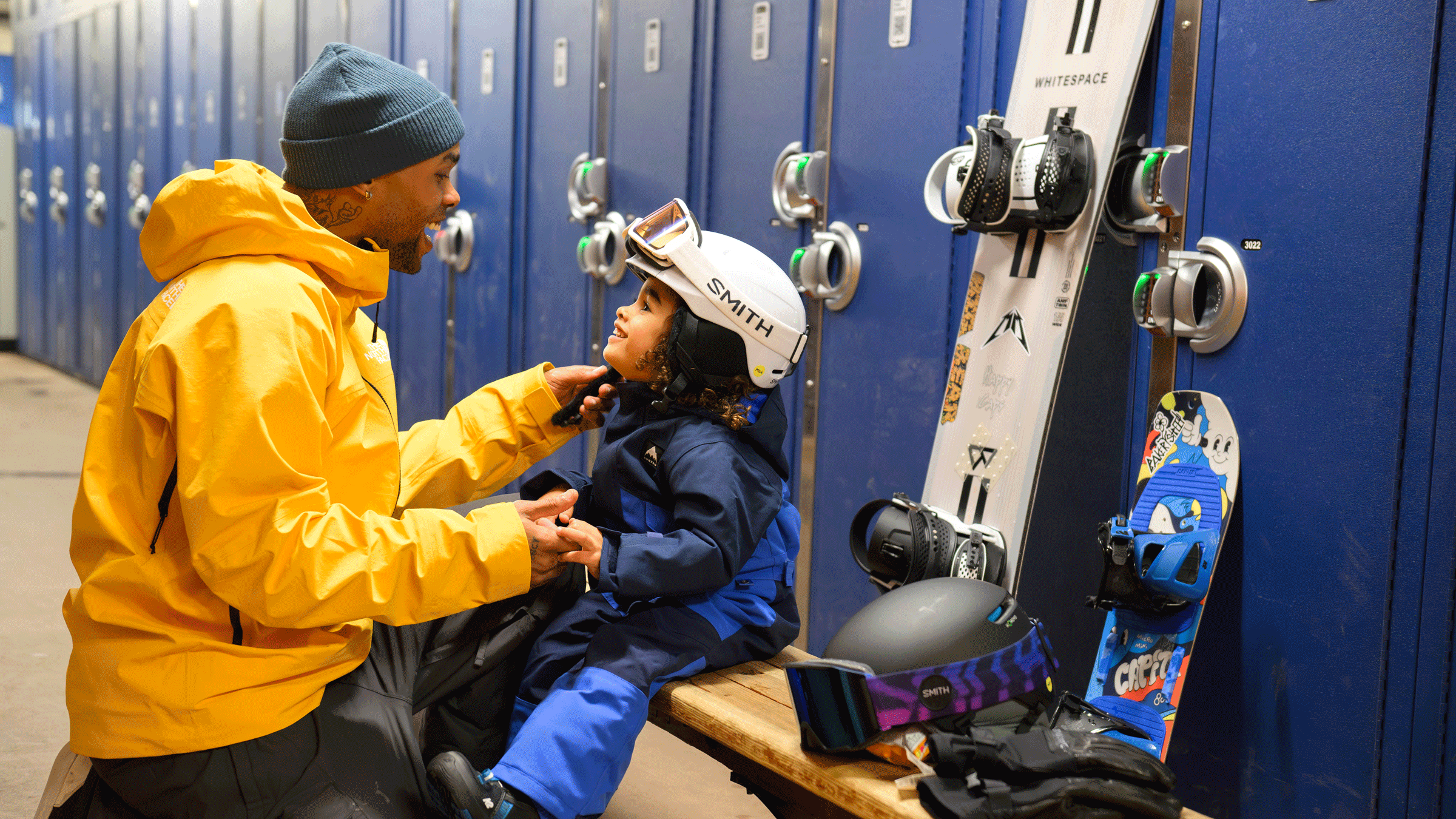 Dad and son sitting in front of tall lockers at a resort with their snowboards leaning behind them in the background.