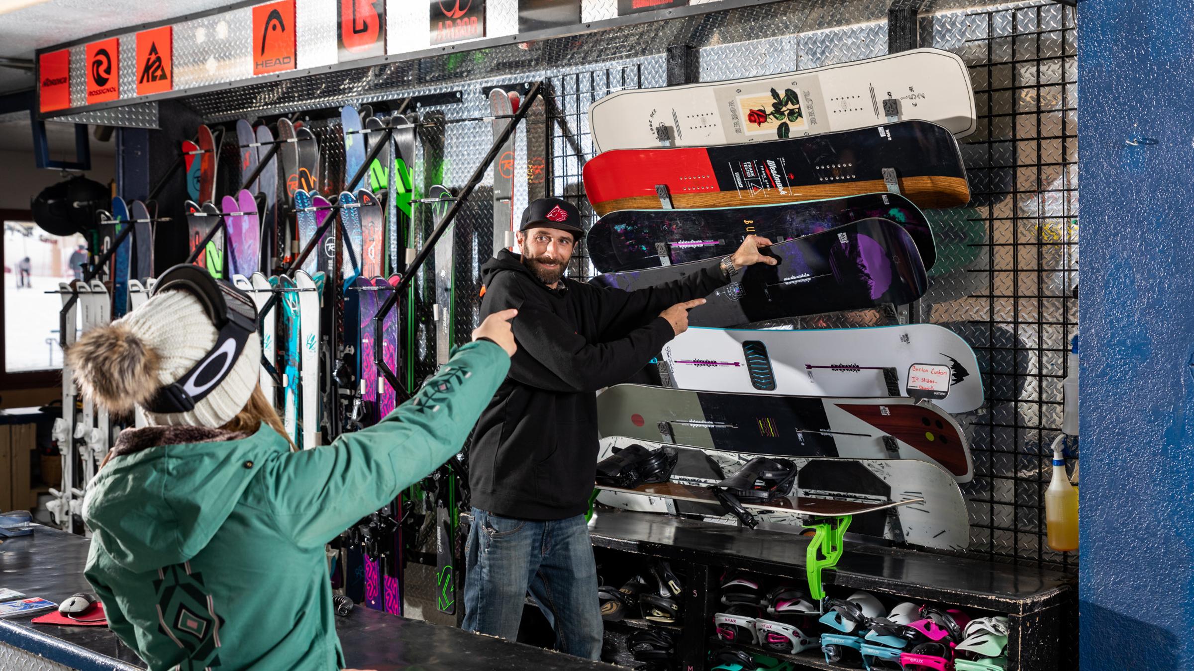 Female in rental shop pointing at which snowboard she wants to rent. 