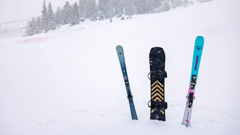 Two skis, both blue in color, and a black snowboard propped into the snow so they are standing straight up on the ski hill during a snowy day.