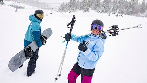 A snowboarder in black helmet and blue jacket holding a grey snowboard standing next to a skier in black with pink pants and blue jacket giving the camera a thumbs up on the slopes during a winter day.