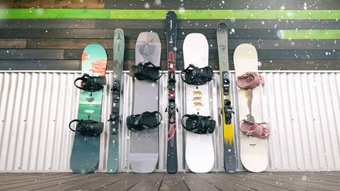 Four snowboard set ups next to three ski set ups, in an alternating pattern , leaned up against a building at Snow Summit with snow falling down from a snowstorm.