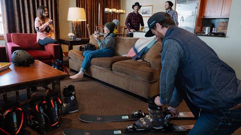 A Ski Butlers employee at a customers lodging accomodation mounting a pair of ski boots to rental skis while four adults lounge in the living room.