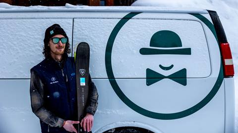 A Ski Butlers employee in sunglasses, a black beanie, flannel shirt, and puffy vest holding a pair of dark colored skis next to a white Ski Butlers rental van.