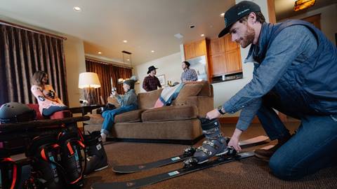 A group of four adults hanging out in their lodging accommodation while a Ski Butlers technician mounts ski boots to a pair of rental skis.