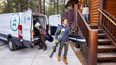 A white cargo van, with the Ski Butlers logo, parked in front of a log cabin and two employees unloading rental equipment to custom fit guests in the comfort of their vacation rental.
