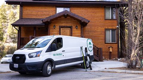 A white cargo van, with the Ski Butlers logo and rental gear leaned against it, parked in front of a wood log cabin in Big Bear Lake.
