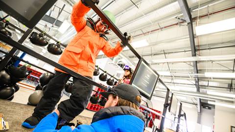A kiddo with a helmet in a orange jacket and black snow pants getting fitted in the Rental Shop with snowboard boots