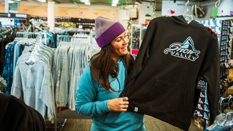 An adult with long hair, wearing a blue sweatshirt and purple beanie, holding up a black long sleeve hoodie sweatshirt with the Snow Valley logo on the front.