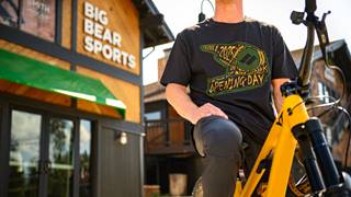 Adult in a black Opening Day Summit Bike Park tee shirt on a orange YT Industries mountain bike in front of the Big Bear Sports retail shop in the base area of Snow Summit.