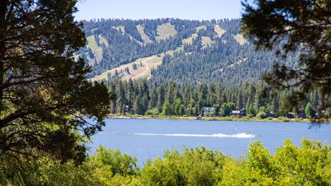 A scenic summer blue sky day from across Big Bear Lake looking at Snow Summit with green forest trees and bushes and a jet ski riding on the blue lake.
