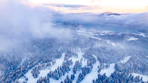 Ariel drone shot of ski resort with sunshine and clouds covering hill and lake in the distance