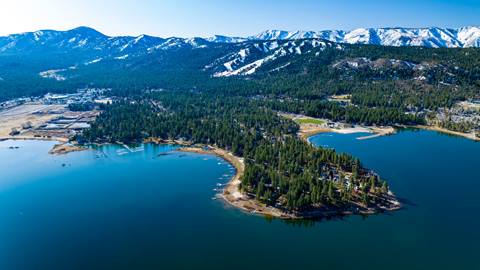 A scenic drone image hovering over Big Bear Lake during the spring time with lush green forest trees and spring mountain peaks with a little snow in the distance.