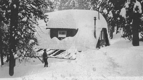 An old black and white photo of an adult standing in powder snow in front of a building that is buried with snow.