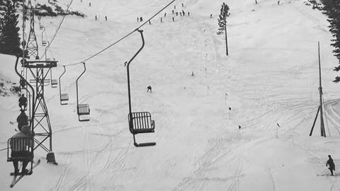 An old black and white photo of an adult sitting on a single chair chairlift with skis on both feet while looking at the wide open ski trail with skiers riding down at Snow Valley.