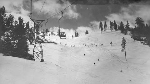 An old black and white photo from a chairlift, on a sunny day with clouds rolling by, while looking at the wide open ski trail with skiers riding down at Snow Valley.