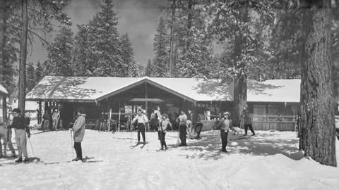 The front of a single story building with skiers in front of the lodge, during a sunny winter day at Fish Camp, now known as Snow Valley.