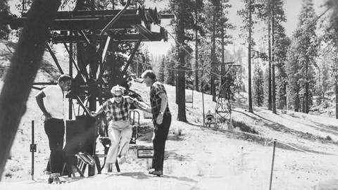 Two adults in dark pants and t-shirts, unloading another adult, wearing light color pants and a checker top, off a chairlift at Fish Camp now known as Snow Valley.