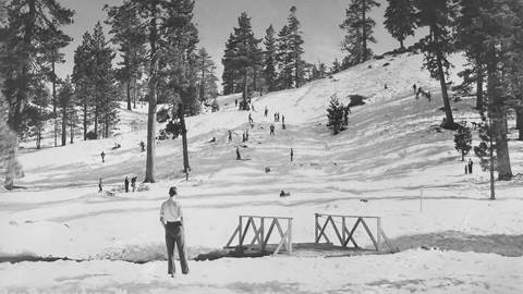 Adult in dark pants and white t-shirt standing near a small wooden bridge at the base of a ski hill, looking up at riders coming down.