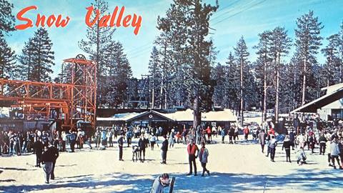 An old color image of Snow Valley with skiers and alpine enthusiasts walking around the base area with snow on the ground, plus on top of building roofs, and blue skies overhead.