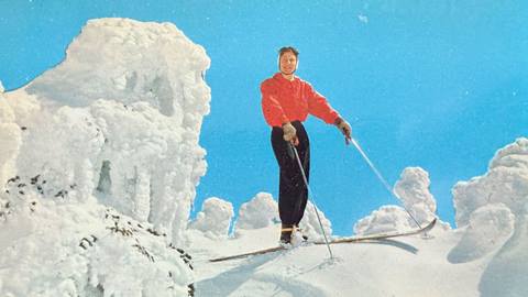 A blue sky day with an abundance of fresh snow at Snow Valley, and a skier in black pants with red long sleeve top smiling for the camera prior to dropping into the trail.