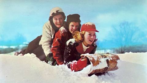Three adults stacked on top of each other laying on their stomachs on a wooden sled that is on fresh powder snow on a blue sky day.