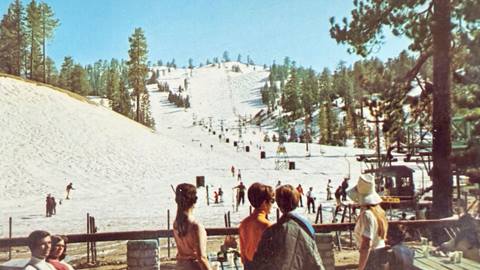 Adults sitting and standing on the Snow Valley sun deck during a sunny winter day, while looking out to the ski hill.