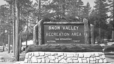 A brick and wood sign that says "Snow Valley Recreation Area" and is placed in the middle of pine trees.