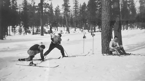 Two adults stretching next to their skis and one adult leaned up against a pine tree near Snow Valley on a winter day.
