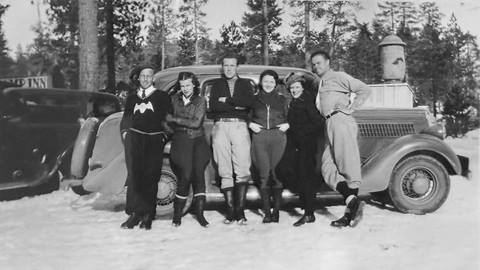 Six adults in long pants, long tops, and boots leaning up against an old car in the snow with pine trees in the background.