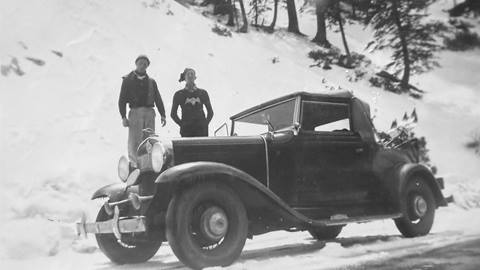 Two adults posing next to their older black vehicle in the snow.