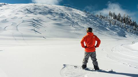 Snowboarder in orange jacket and helmet looking up at Slide Peak from the base of the peak