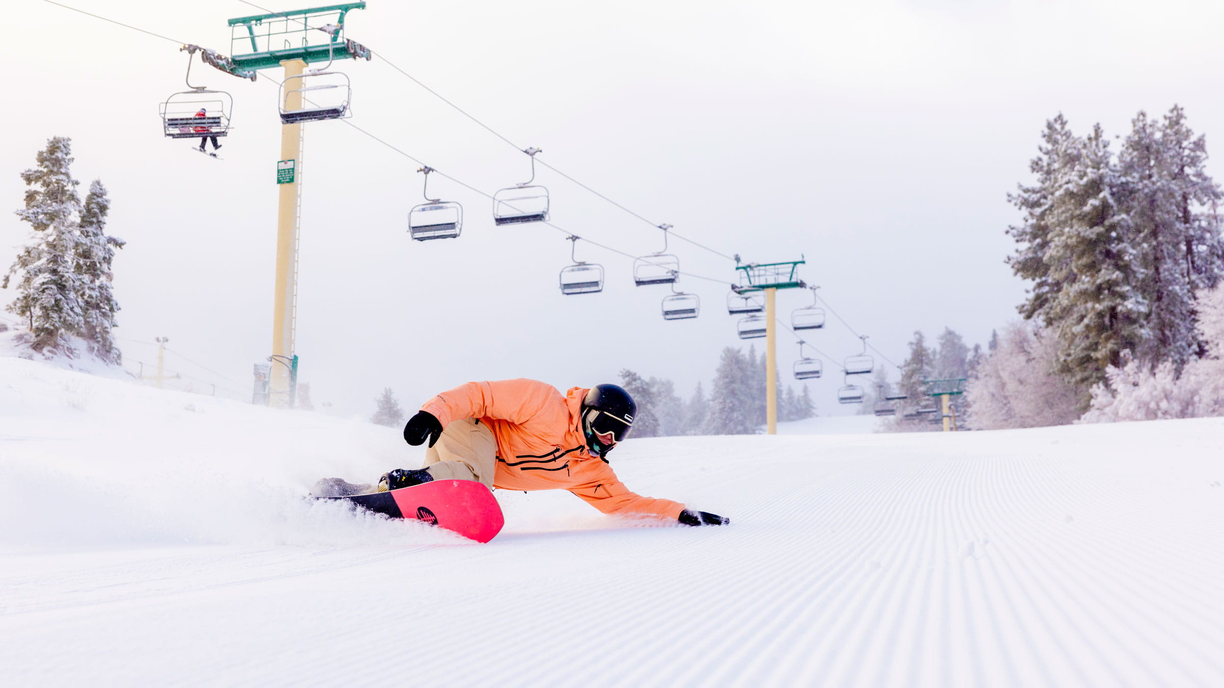 Snowboarder in orange jacket and black helmet carving corduroy snow with left arm dragging on snow.