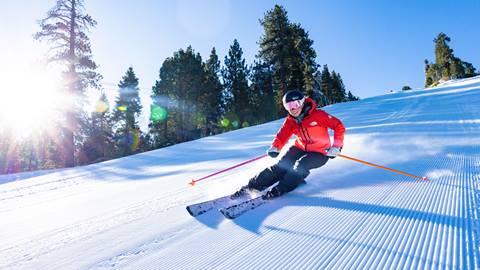A skier wearing black pants, a red jacket, white snow goggles, and a black helmet, riding down a freshly groomed corduroy ski trail at Snow Summit on a sunny blue sky winter morning.