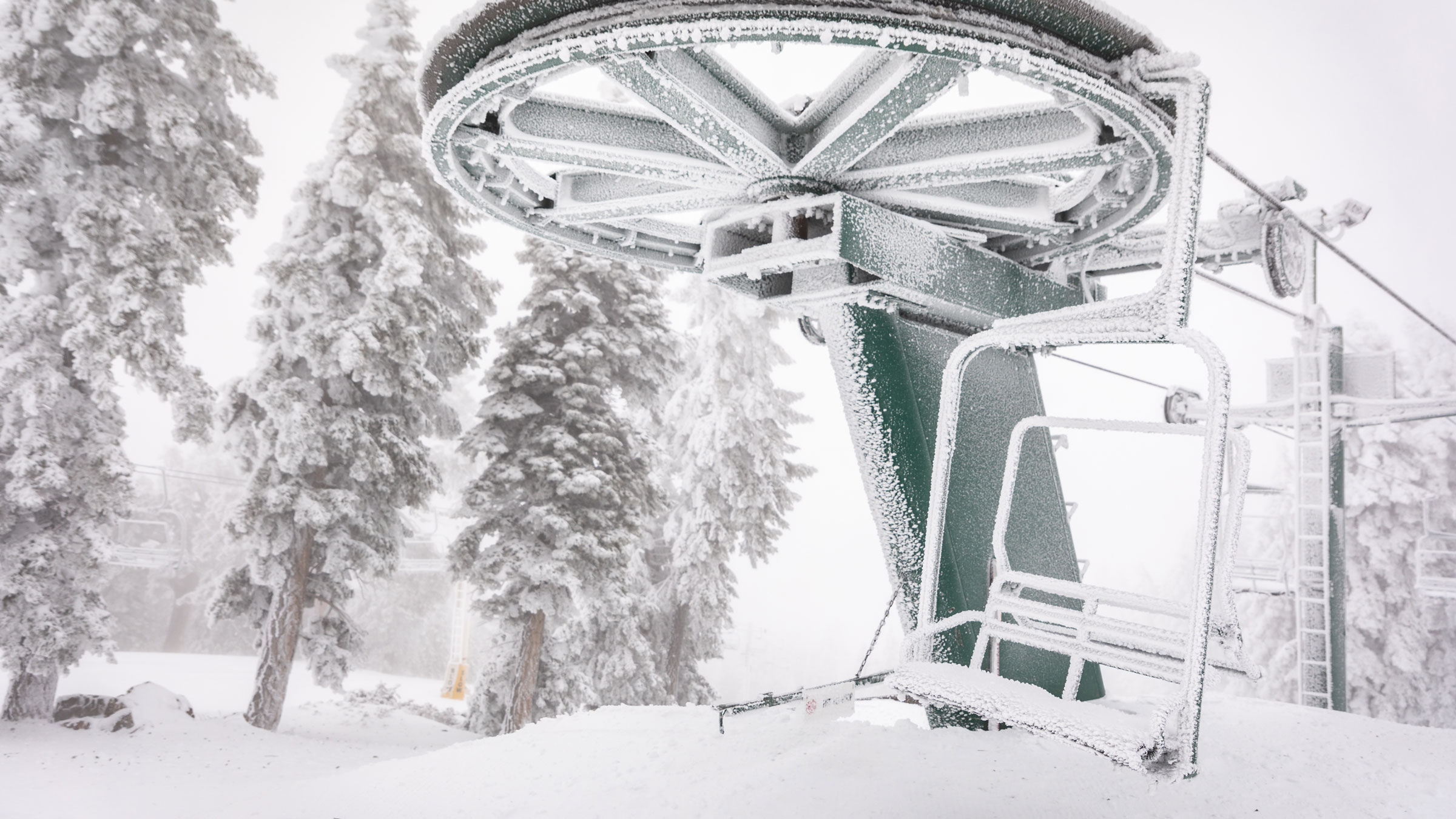 A view from the top of a Big Bear Mountain Resort chairlift, during a stormy winter day, with the chairlift buried under fresh snow and the trees in the background covered with a white blanket of snow.