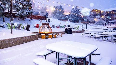 Snow Summit's base area with a fresh coat of snow on the walkways, fire pit, tables, and chairs during a snowstorm and the string lights turned on.