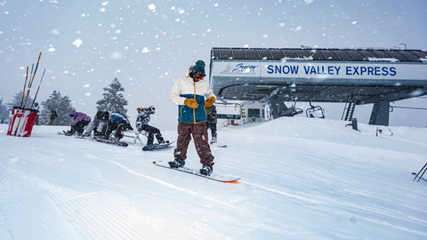 A snowy storm day from the top of Snow Valley's Chair 1 with skiers and snowboarders strapping into their setups in preparation of heading down the powdery trails.