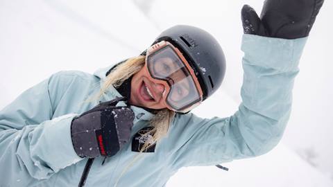 Adult in blue jacket, snow goggles, and helmet with one arm raised smiling at camera