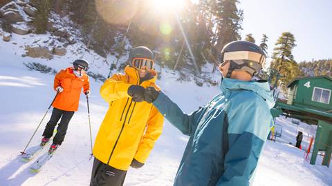 Three adults, two of them fist bumping eachother, in colorful winter snow jackets, waiting at the bottom of a chairlift line at Bear Mountain on a sunny day during winter time.