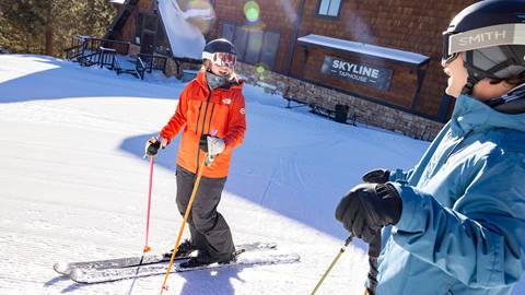 Two skiers wearing black helmets, one in a blue snow jacket with grey Smith goggles and the other in a red jacket with white snow goggles, mounted on their skis at the top of Snow Summit on a sunny winter day outside Skyline Taphouse food venue.