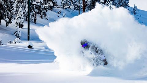 Snowboarder in black helmet and reflective goggles slashing through powder 