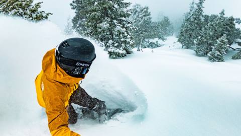 A snowboarder in a yellow jacket and black helmet with black Smith goggles riding through a trail with trees and fresh snow.