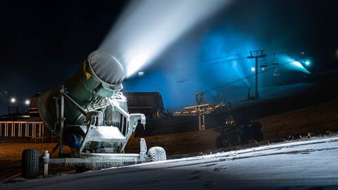 A snow gun blowing snow during the nightfall at Bear Mountain with additional snow guns activated and blowing snow in the background on a separate trail.