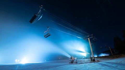 An image looking up towards black starry night sky with a chairlift, lights, and snow making guns turned on making snow for the mountain trails.