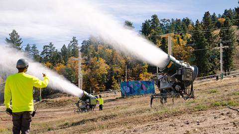 Adult in a dark hardhat and bright yellow long sleeve shirt giving the OK to the on-hill snowmaking crew while they test two new snow machine fan guns during a sunny fall day. 