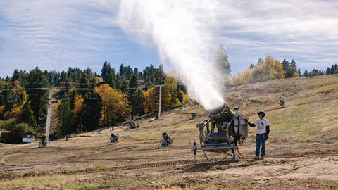 Adult in a dark hardhat and white shirt standing next to an on-hill snowmaking fan gun machine that is turned on during a sunny fall day. 
